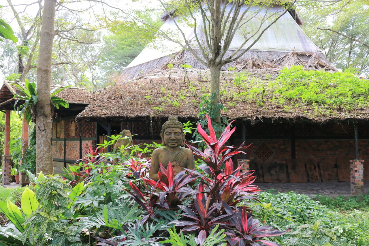 A tranquil garden featuring a Buddha statue amidst vibrant plants and rustic architecture.