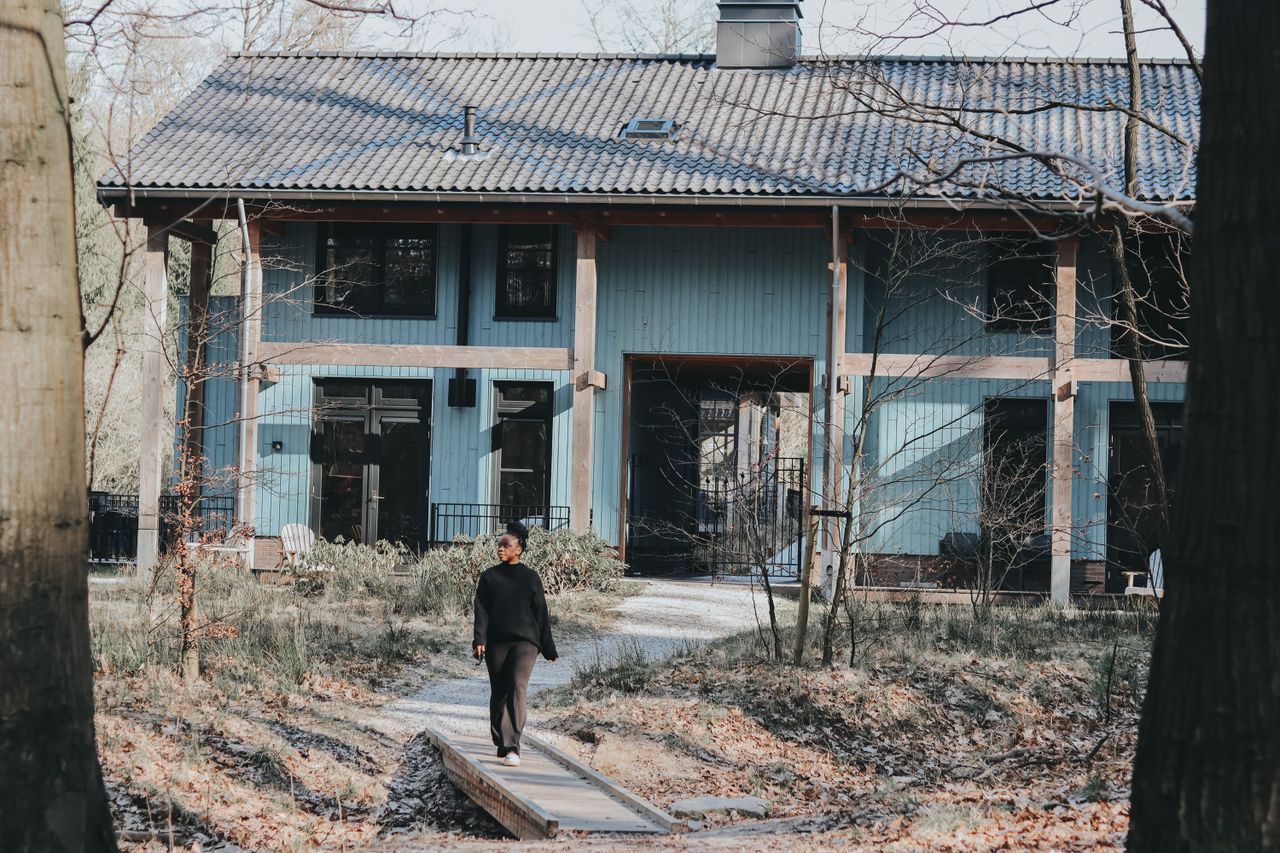 A woman walks on a footbridge toward a modern house in a wooded rural area in the Netherlands.
